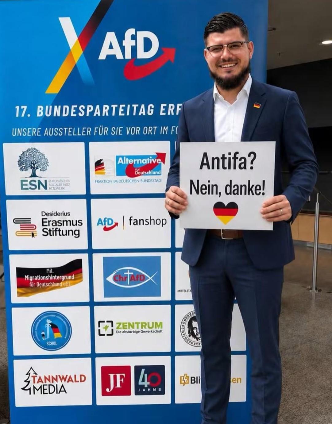Ein dunkelhaariger Mann mit Brille und Bart im Anzug mit weißem Hemd. Er hält ein Schild mit der Aufschrift Antifa? Nein, danke! Und Herz in Deutschlandfarben.
Er steht neben einem blauen Aufsteller, 17. Bundesparteitag Erfurt. Darauf sind mehrere rechte und rechtsextreme Unterstützer*innen bzw Teilnehmer*innen des Parteitages, zb Erasmus Stiftung, es, jf, tannwald Media AFD Fanshop