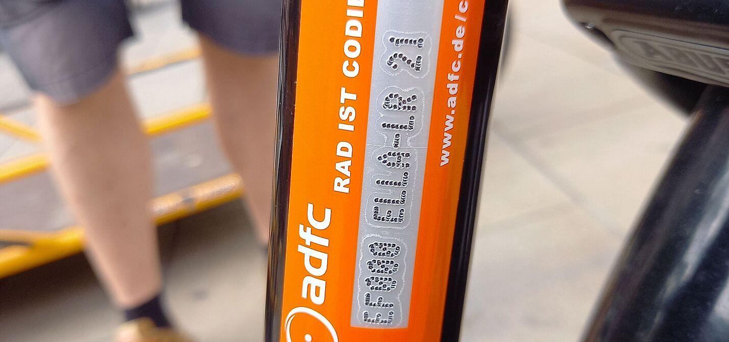 Muster einer Codierung. Orangenes Rahmenrohr mit silbernem Streifen, darauf gestichelt ein Code aus 13 Buchstaben und Zahlen.