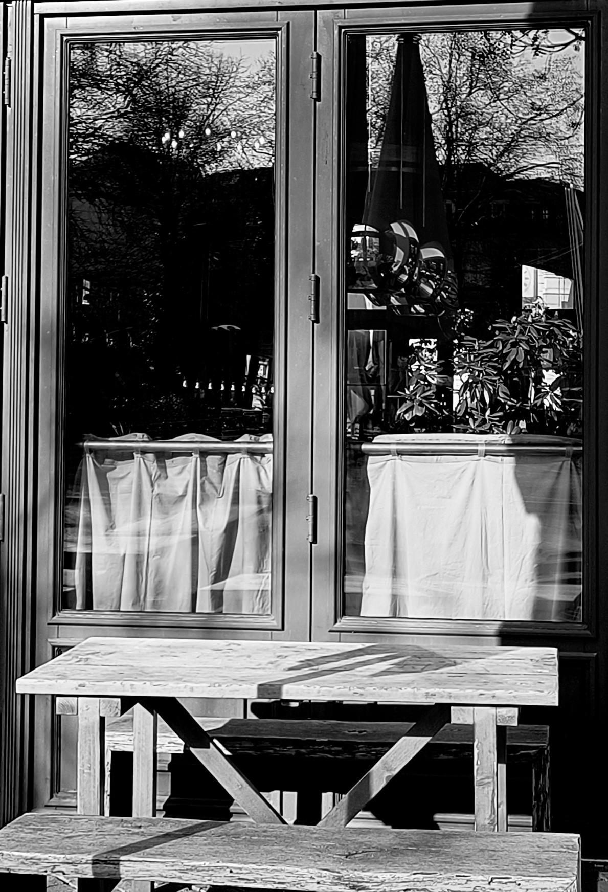 Ein Schwarzweiß-Bild eines hohen, rechteckigen Fensters mit Bistro-Gardinen im unteren Drittel, von außen fotografiert. Draußen davor steht ein Holztisch mit zwei Bänken. 

A black-and-white photograph of a tall, two-part window with bistro curtains in the lower third, taken from the outside. Outside, in front of it, stands a wooden table with two benches.