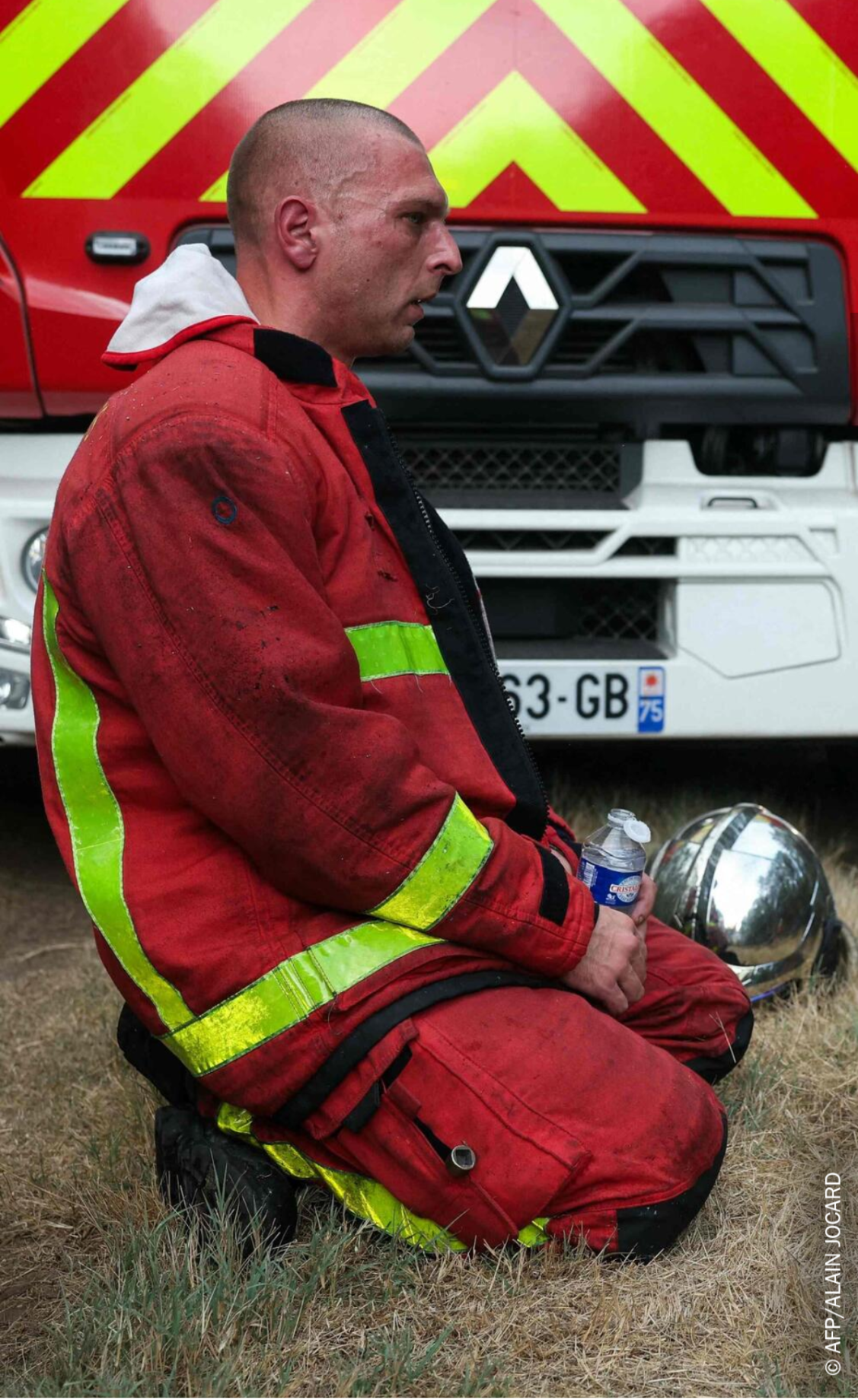 Un pompier aux cheveux coupés court, le visage rougi par la fatigue, est agenouillé sur l'herbe sèche devant un camion de pompiers, vêtu d'une tenue de travail tachée de rouille. Son casque est posé à côté de lui ; il tient dans ses mains une bouteille en plastique remplie d'eau. 

A firefighter with closely cropped hair and an exhausted, flushed face is kneeling on the dry grass in front of a fire engine, wearing rusty work clothes. His helmet lies beside him; in his hands he is holding a plastic bottle of water.