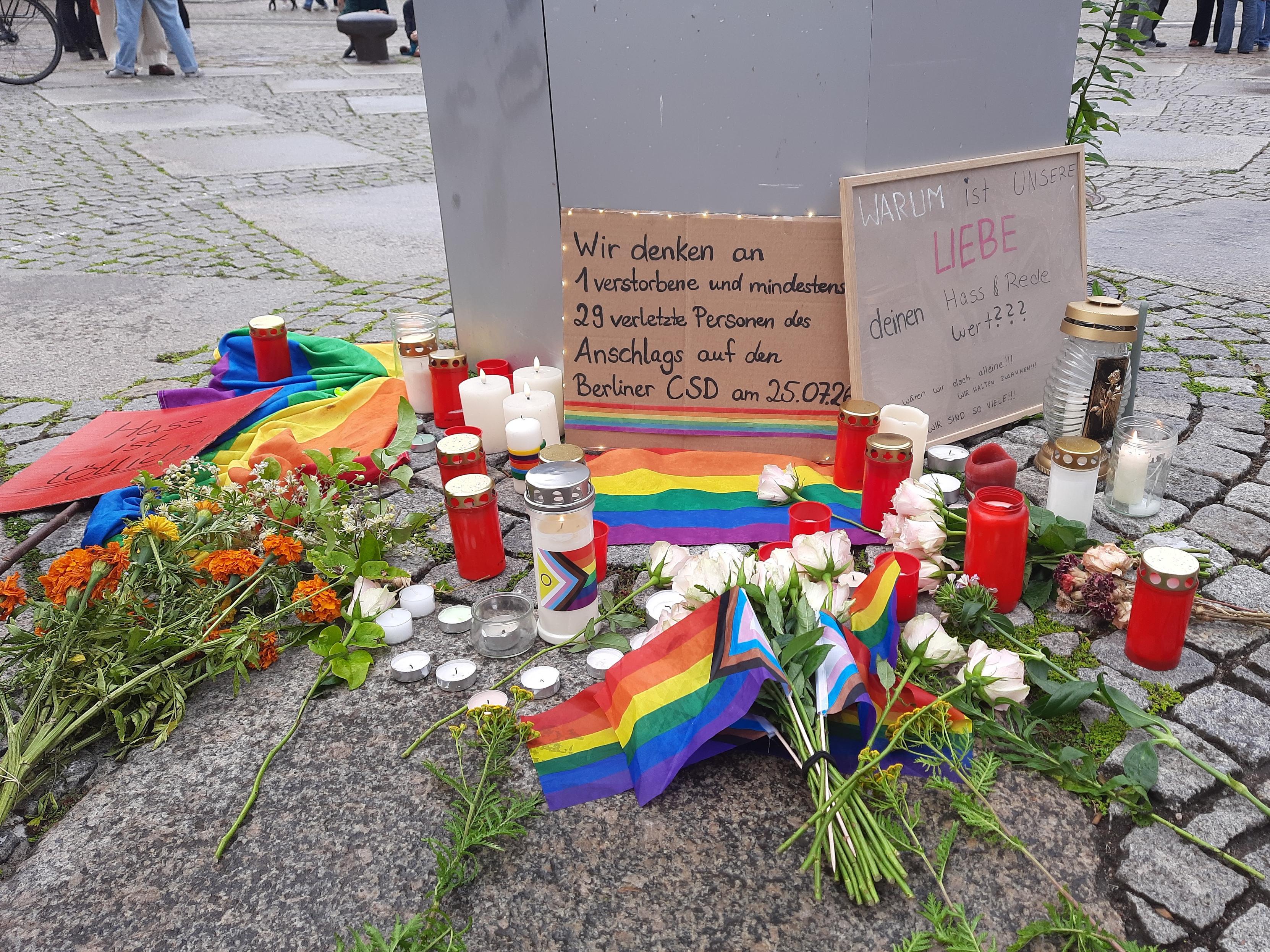 Auf dem gepflasterten Boden liegen Blumen, Blumensträuße, Prideflaggen, Kerzen. Im Hintergrund lehnen an einer Säule zwei Schilder.
Linkes Schild, mit Lichterkette umrandet: Wir denken an 1 verstorbene und mindestens 29 verletzte Personen des Amschlags auf den Berliner CSD am 25.07.2026.
Rechtes Schild: Warum ist unsere Liebe deinen Hass & Rede wert??? Darunter kleiner geschrieben, wären wir doch alleine!!! Wir halten zusammen!!! Wir sind  so viele!!!
