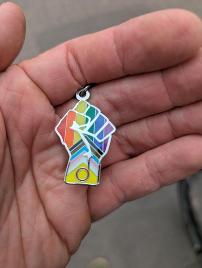 Foto eine Schlüsselanhängers in einer offenen hand. Der Metall-Anhänger zeigt eine in die Luft gereckte Faust. Die Faust ist mit der progressive pride flag gefüllt.