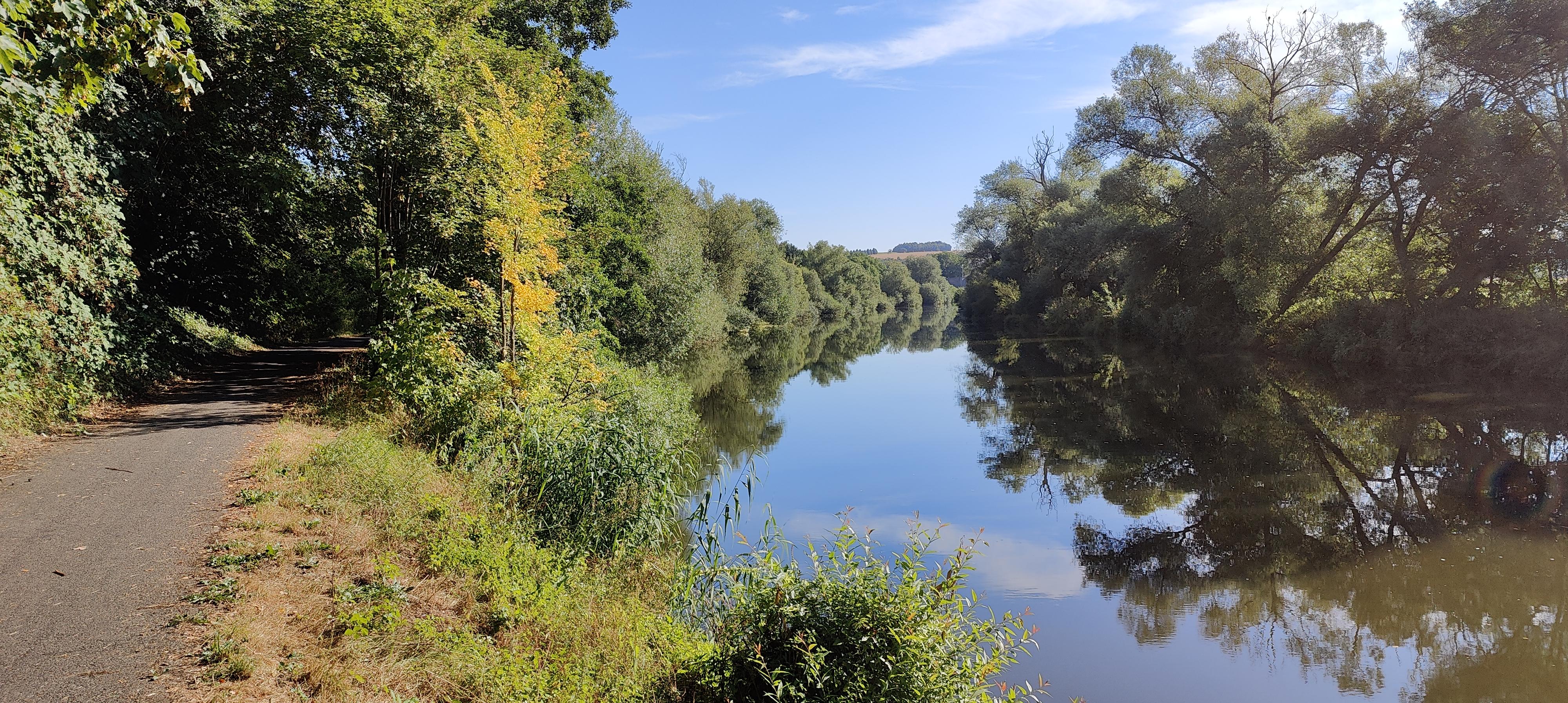 Blick auf die Lahn, die umsäumt ist von Bäumen, welche sich auf der Wasseroberfläche spiegeln. Der Himmel ist blau