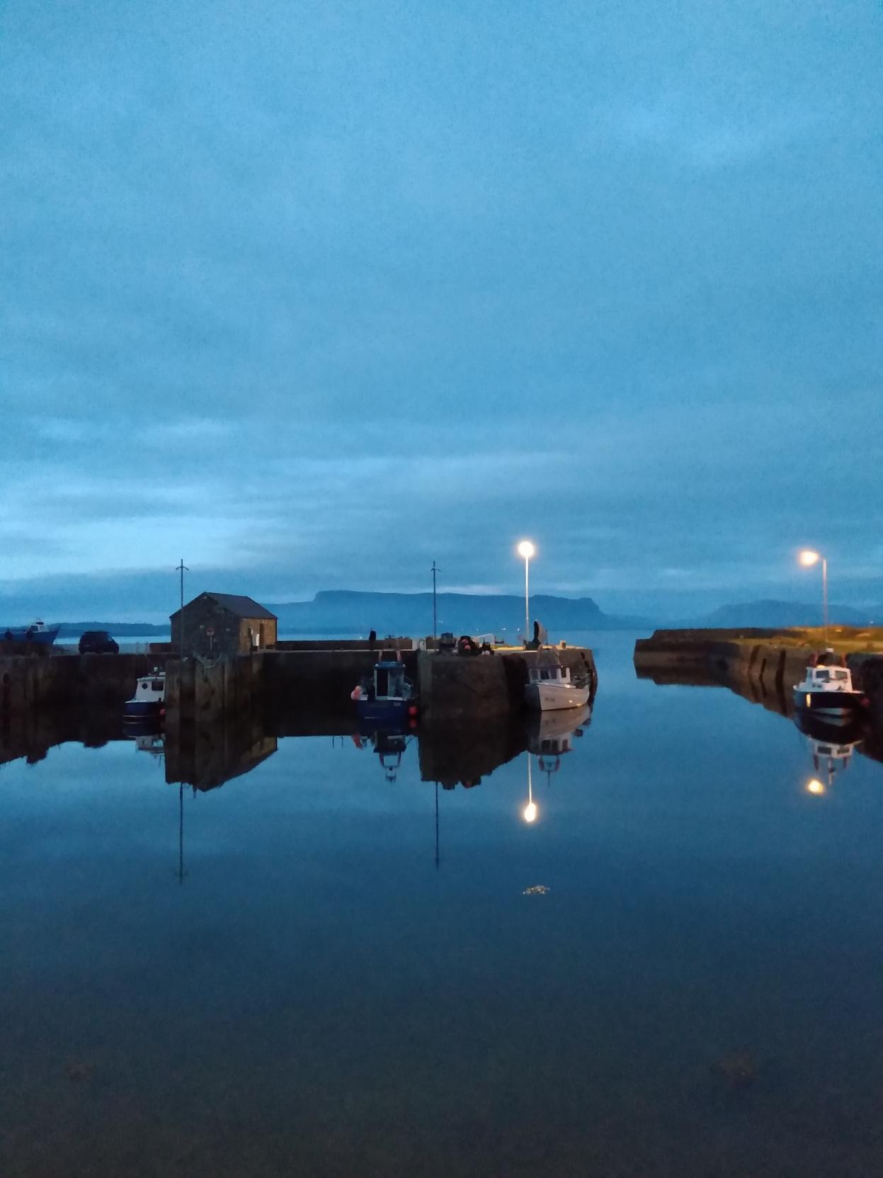 A small, old harbour with stone quay walls and a few small boats lies peacefully in the blue light of the falling night. The waters of the Atlantic, both inside and outside the harbour walls, are unusually calm and as smooth as glass. The light from two small lanterns is reflected in it like tiny dots. The sky and the water are both a blurred dark blue, and on the horizon, the striking silhouette of the table mountain Benbulben looms in the distance. 

Ein kleiner alter Hafen nit Kaimauern aus Stein und einzelnen kleinen Booten liegt ruhig im blauen Licht der einbrechenden Nacht. Das Wasser des Atlantik ist innerhalb und außerhalb der Hafenmauern ungewöhnlich ruhig und spiegelglatt. Das Licht von zwei kleinen Laternen spiegelt sich wie kleine Punkte darin. Himmel und Wasser sind gleichermaßen verschwommen dunkelblau, und am Horizont zeichnet sich in der Ferne die markante Silhouette des Tafelbergs Benbulben ab.