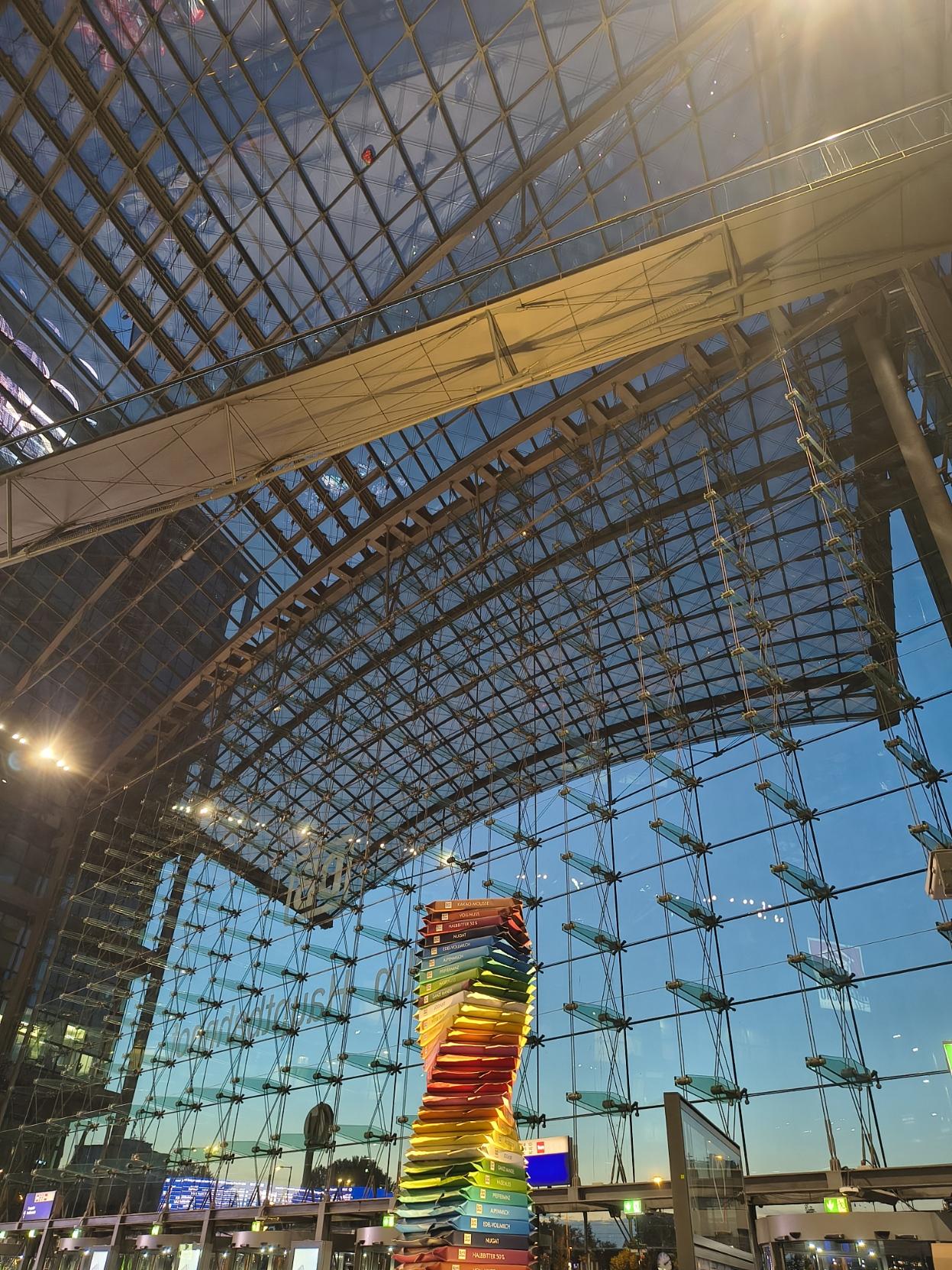 The impressive, soaring glass-roofed vault of Berlin Central Station, with its vast façade of windows. Outside, the light takes on a blue hue as night falls; inside, in the entrance hall, a huge, colourful tower has been erected from square, twisted, rainbow-coloured, oversized chocolate bars. 

Das beeindruckende hohe verglaste Gewölbe des Berliner Hauptbahnhofs mit der riesigen Frontseite aus Fenstern. Draußen scheint das Licht blau von der hereinbrechenden Nacht, innen ist im Eingangsbereich ein riesiger bunter Turm aus viereckigen verdrehten  regenbogenfarbigen überdimensionierten Schokaladentafeln aufgebaut worden.