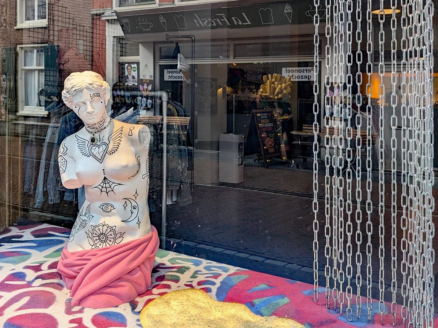 Man sieht in ein Schaufenster. 
Der Fokus liegt links auf einer dezent grafisch bemalten, ansonsten weißen, Büste. Diese steht auf einem rot/blau/weiß gemusterten Teppich.
Hinter der Büste spiegeln sich die unterschiedlichsten Dinge: eine Hauswand mit Fenster, ein Kleiderständer, eine Tüte mit übergroßen Plastikpommes etc. 
Rechts hängt von oben bis unten eine Art Kettenvorhang.