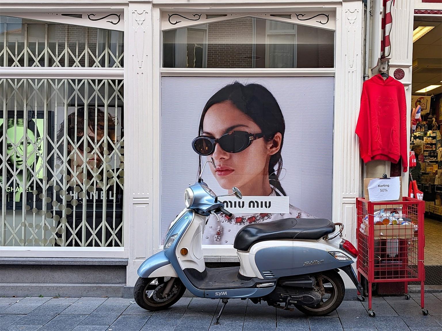 Eine weiß-blaue Vespa (oder ähnlich) steht vor einer Hauswand. 
Links, etwa zu einem Drittel ein weißes Gitter. Daran schließt mittig ein Plakat an, auf dem eine junge, dunkelhaarige Frau mit Sonnenbrille zu sehen ist. Ein Werbetext auf Halshöhe: "miu miu". Der Roller steht genau vor diesem Plakat.
Ganz rechts der Eingang zu einem Laden. Neben dem Plakat hängt ein roter Pullover.