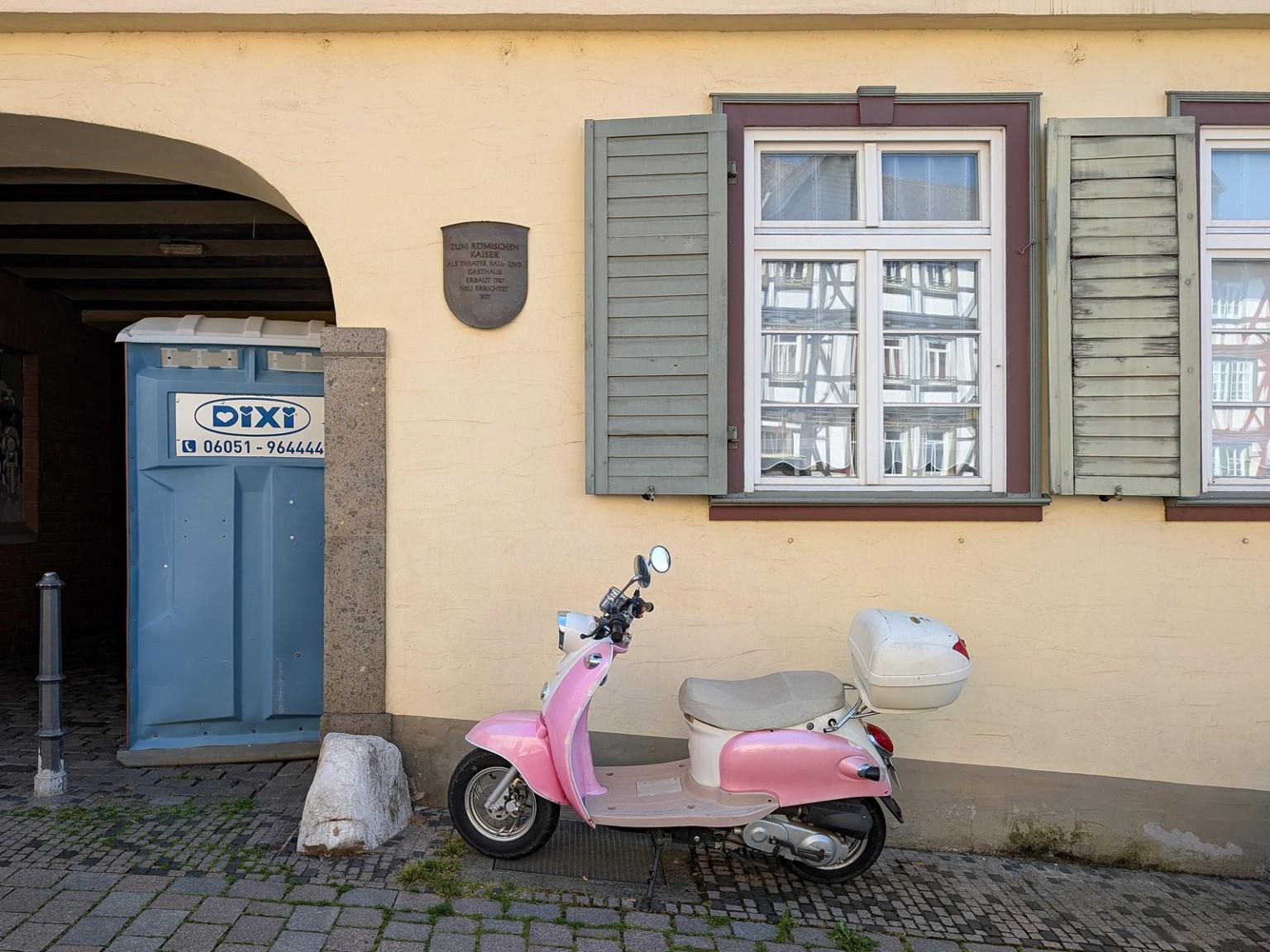 Gelbe Hauswand
Links in einem Torbogen steht ein blaues Dixi-Klo. 
In der Hauswand ist ein Fenster mit geteilten Scheiben, in denen sich ein Fachwerkhaus spiegelt, und grauen Fensterläden.
Vor all dem steht ein bonbonrosa-farbener Motorroller