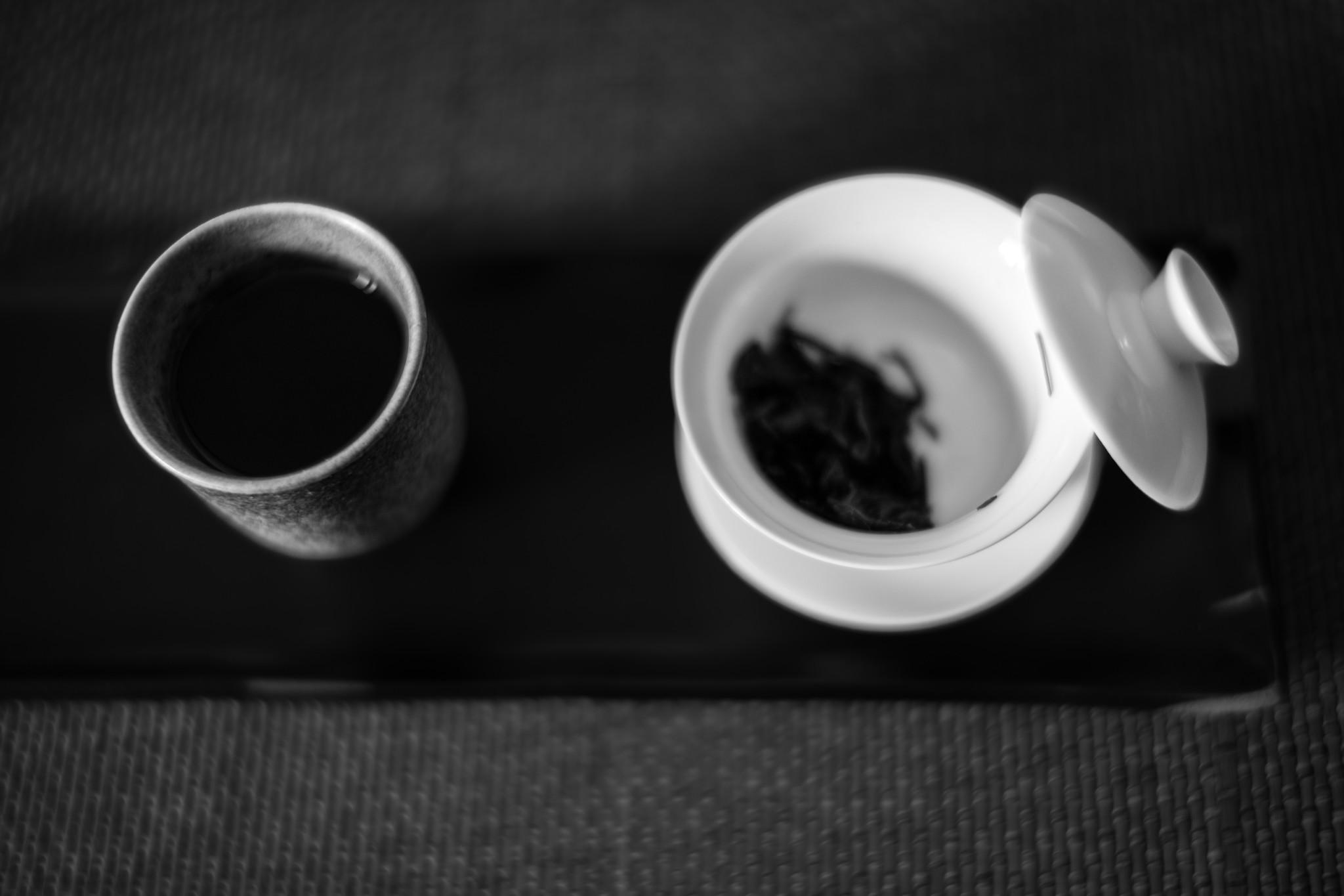 Schwarzweißaufnahme von oben: ein weißer Porzellan-Gaiwan mit trockenen, dunklen Teeblättern, der Deckel liegt schräg auf dem Rand. Links daneben eine dunkle, gesprenkelte Keramiktasse mit aufgegossenem Tee, beides auf einem dunklen Teeboot auf einer Bambusunterlage.