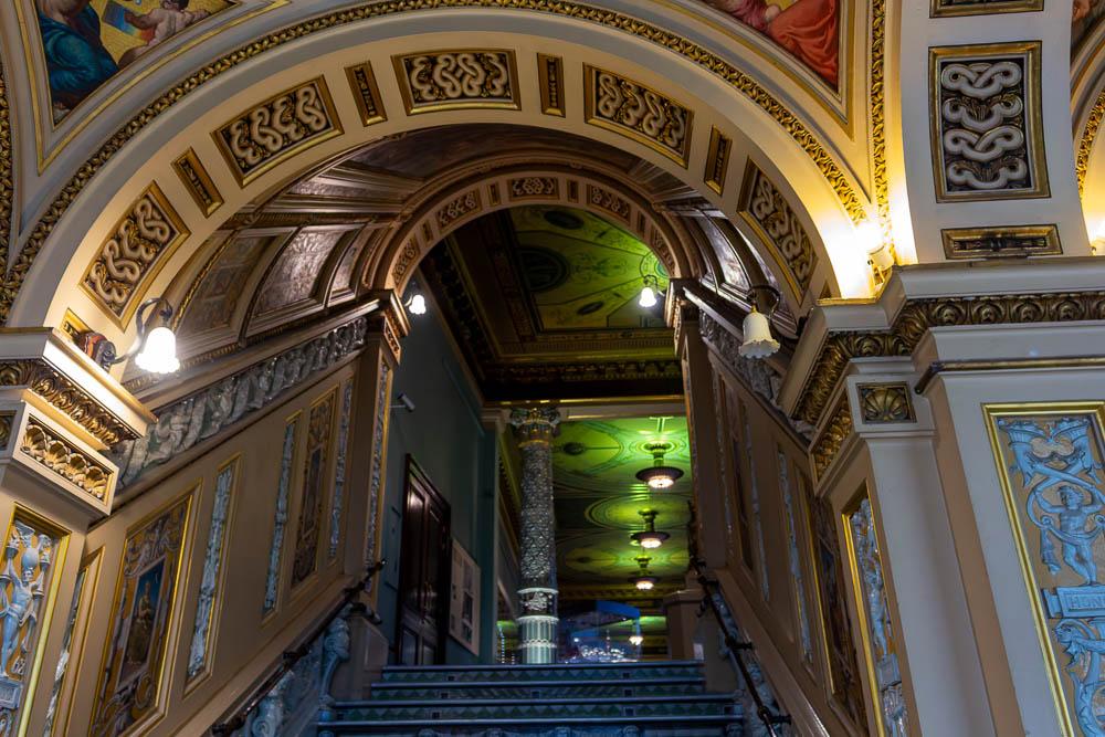 Aufwendig dekorierter Treppenaufgang im Victoria and Albert Museum in London. Ornamente, Wandbilder und eine reich verzierte Decke rahmen die Treppe, die in einen weiteren Ausstellungsraum führt.