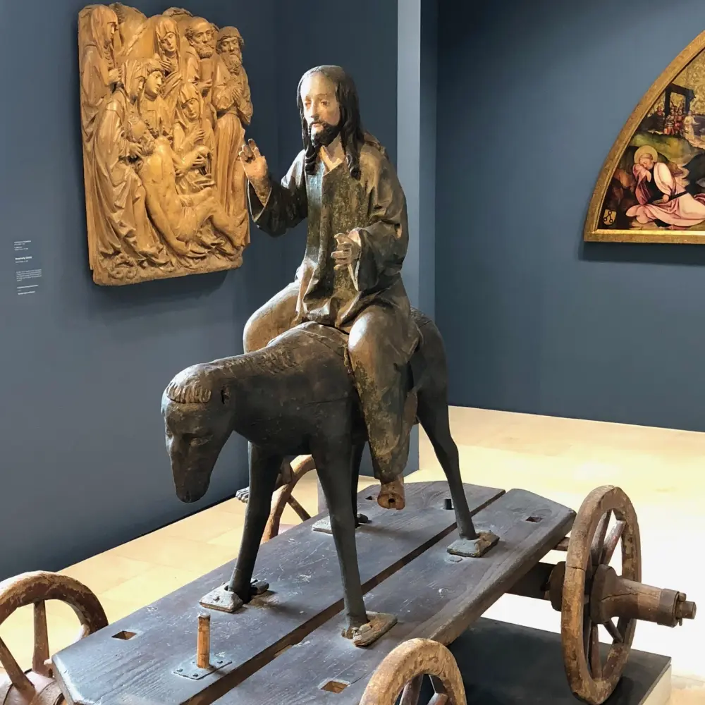 In der Mitte ein Esel mit Reiter auf einem hölzernen Wagen; links oben ein Holzrelief; rechts oben Teil eines alten Ölgemäldes