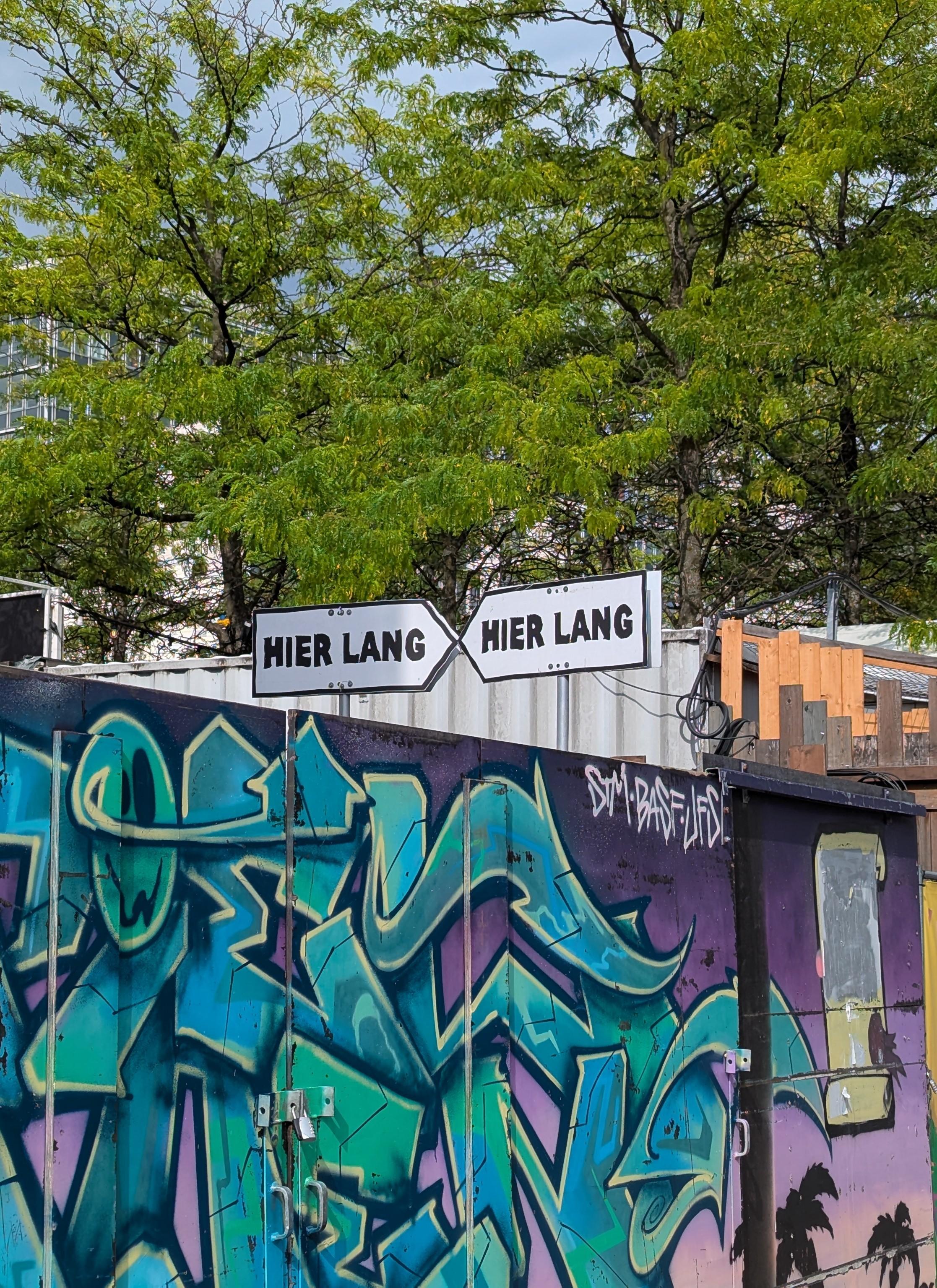 Zwei Schilder mir der Aufschrift "Hier lang" zeigen genau aufeinander. Sie stehen über einer mit Graffiti verzierten Wand.