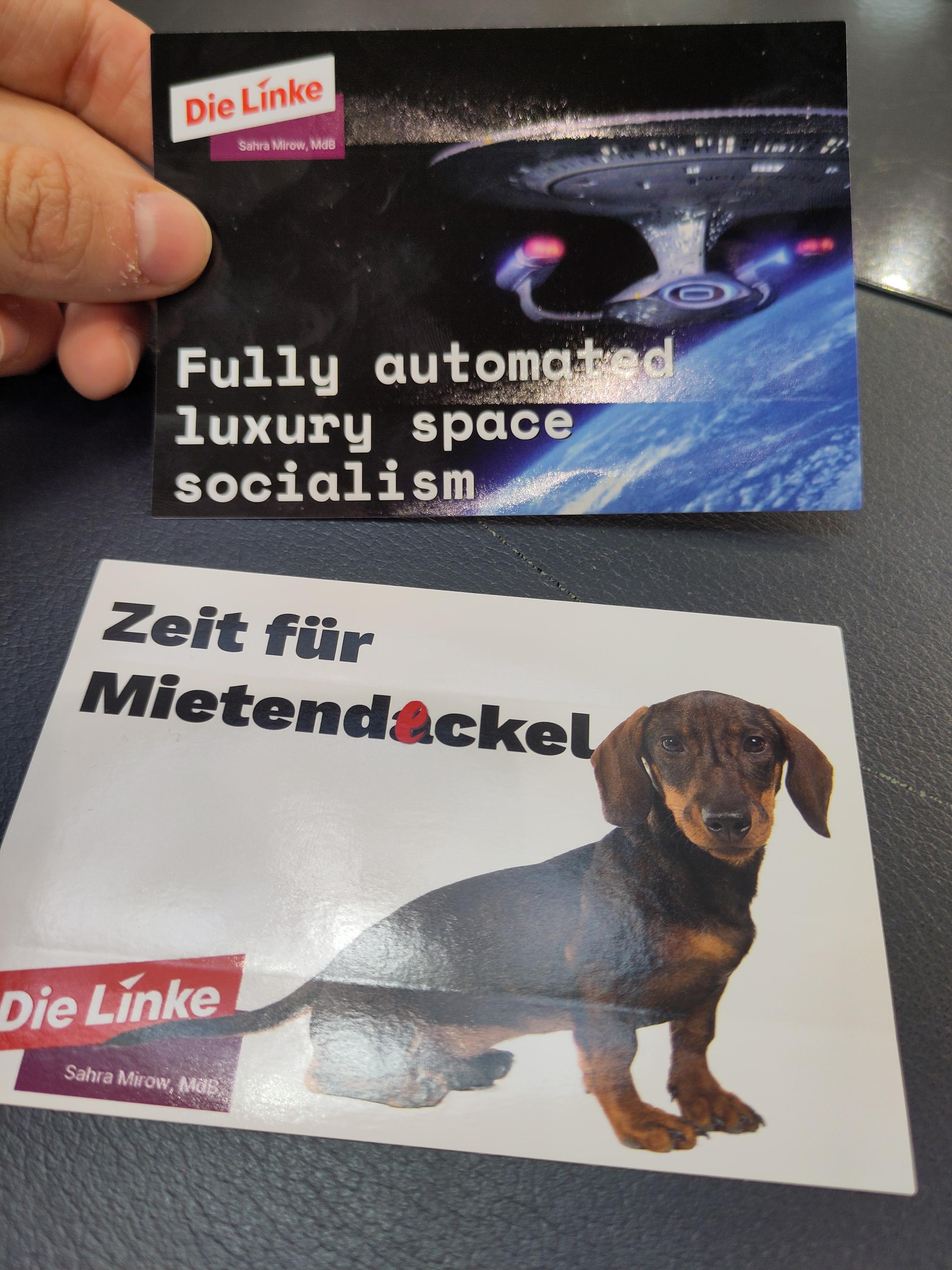 Zwei Sticker liegen auf einem Tisch. Auf dem ersten ist eine Star Trek Raumschiff mit den Text "fully Automated space socialism", auf dem zweiten ein niedlicher Dackel mit dem Text Zeit für Mietendackel /deckel. Beide sind von Sarah Mirow 
