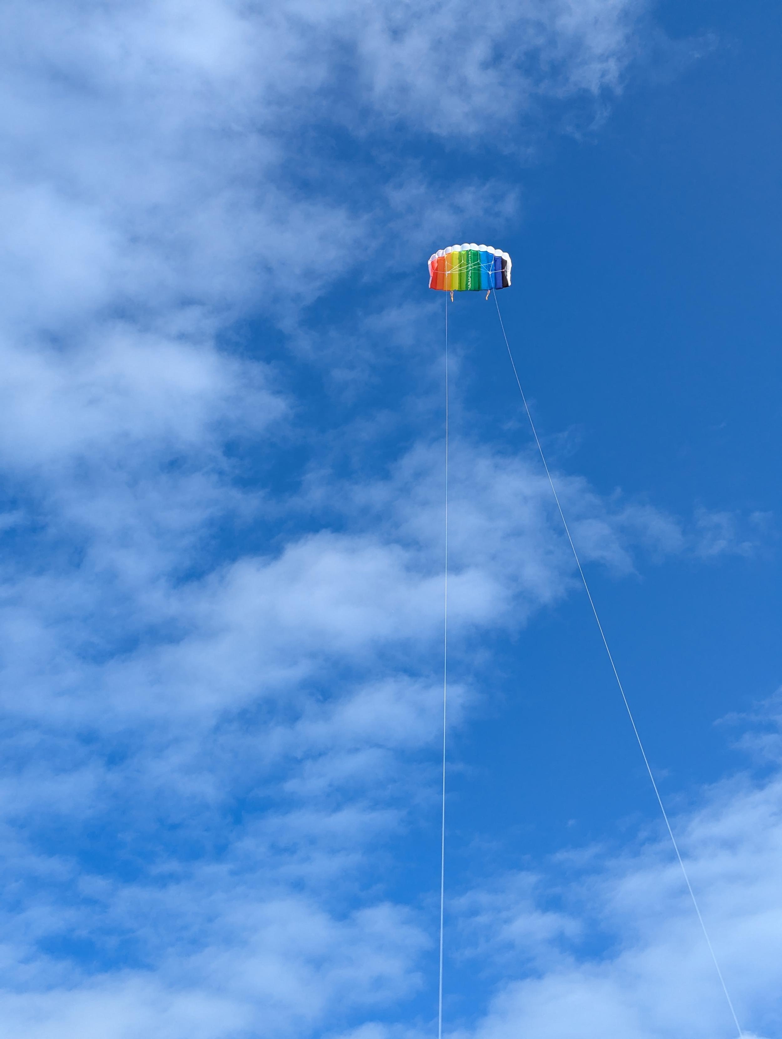 Ein bunt gestreifter Flugdrachen schwebt hoch am Himmel. Zwei lange Schnüre reichen vom Drachen nach unten aus dem Bild. Der blaue Himmel ist von weißen, locker verteilten Wolken durchzogen.