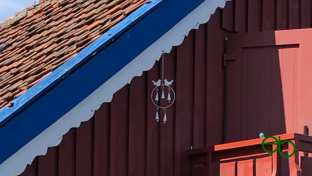 White wind chime with two birds sitting on a ring with five bells hanging at the roof of a red wooden cottage.